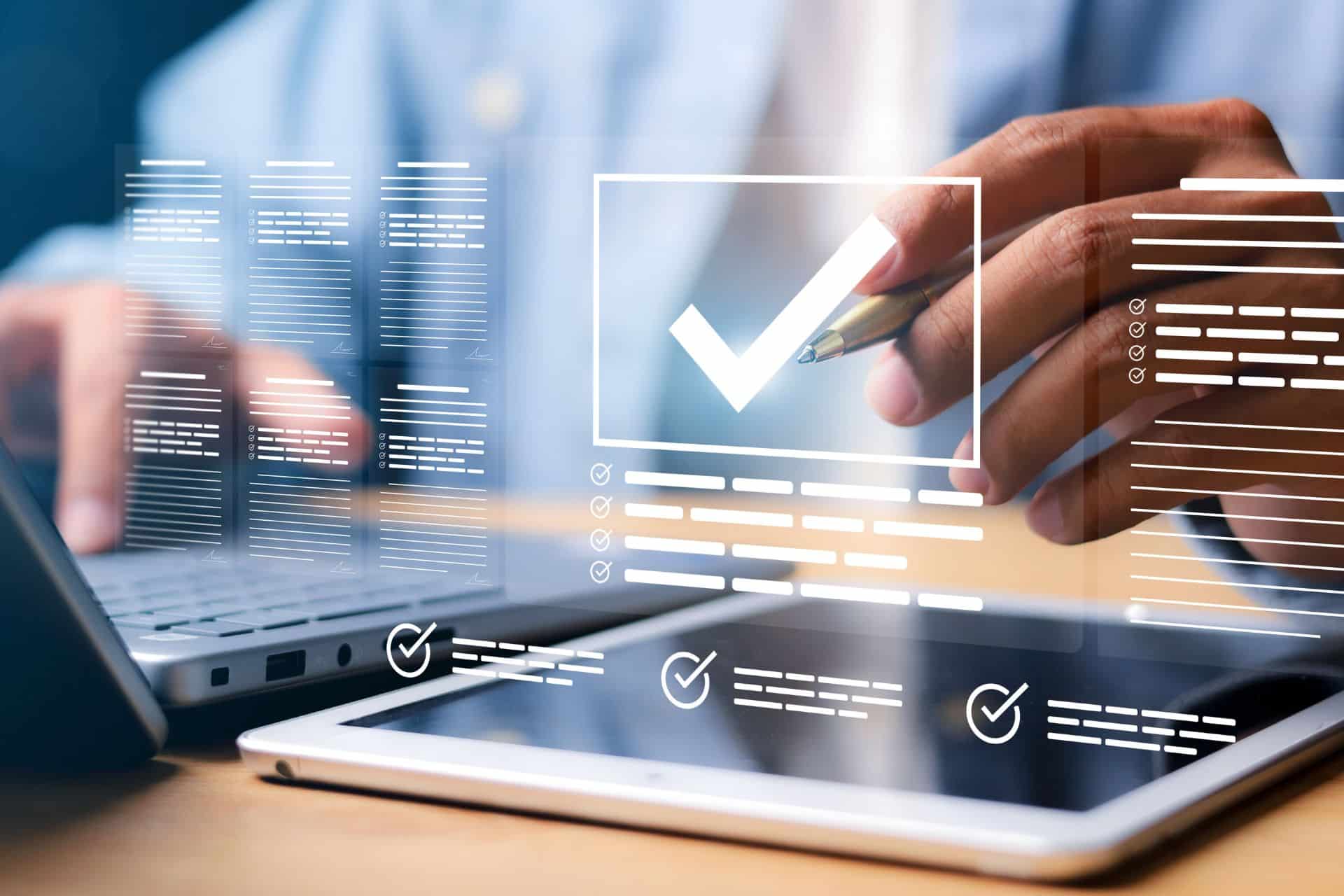 Main tenant un stylo devant un ordinateur et une tablette affichant des checklists pour un cahier des charges informatique